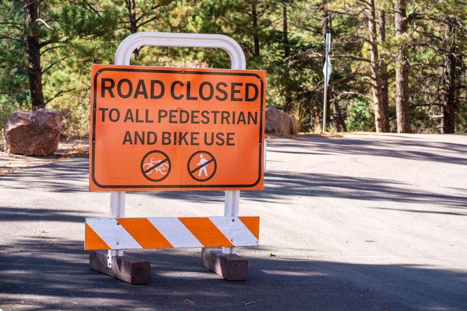A rectangular orange road closure sign with black text is positioned on a paved street, indicating that the road is closed to all pedestrian and bicycle use. The sign is mounted on a white metal frame