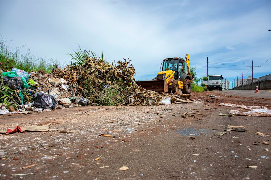 A scene showing a large pile of mixed waste and recyclables, including green plastic bags, black rubbish bags, cardboard, and organic debris, on the side of a dirt road. A yellow backhoe loader with a front bucket attachment is actively moving the waste pile, with its cab facing towards the viewer. The area appears to be in an outdoor urban or suburban environment, with a partly cloudy sky overhead and a row of buildings, parked vehicles, and utility poles visible in the background. The ground around the waste shows scattered smaller pieces of debris and paper, indicating ongoing cleanup or disposal activity. This image illustrates a home relocation or waste collection process that aligns with the recycling rules outlined by Barnet Council, which Man with Van Woodside Park assists with during house removals and furniture transport.
