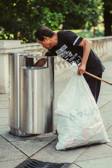 A row of four recycling bins positioned on a paved area outside a property, designated for different waste materials including plastic, glass, paper, and metal, each with distinct colours and clear labels in Cyrillic script. The red bin for plastic features a lid slightly open, with a small portion of plastic waste visible. The green bin for glass is closed, while the blue bin for paper and the yellow bin for metal are both sealed. Behind the bins, there is a lush green landscape with well-maintained grass, a small evergreen tree, and other trees with dense foliage. To the left of the bins, a bicycle rack with a curved metal frame is visible. In the background, outdoor furniture such as orange sun loungers and a red picnic table, along with a row of parked bicycles, are set on a grassy area under the shade of large trees. The image is well-lit with natural daylight, and the scene captures a typical outdoor setting related to home relocation and waste management during a move, as managed by Man with Van Woodside Park during packing and moving processes near Woodside Park.