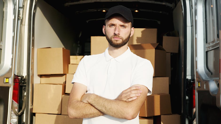 A man with a serious expression, wearing a white polo shirt and black cap, stands with arms crossed inside an open commercial van used for house removals. Behind him, the van is loaded with numerous cardboard boxes, some sealed with packing tape, arranged on the van's floor and against its walls. The interior of the van is well-lit, showing a tidy loading area with packed boxes ready for transportation. The scene captures part of the vehicle’s interior and the outside pavement visible at the edges of the van’s opening, reflecting a professional home relocation process handled by Man with Van Woodside Park, supporting efficient furniture transport and packing services for House removals in Woodside Park N12.