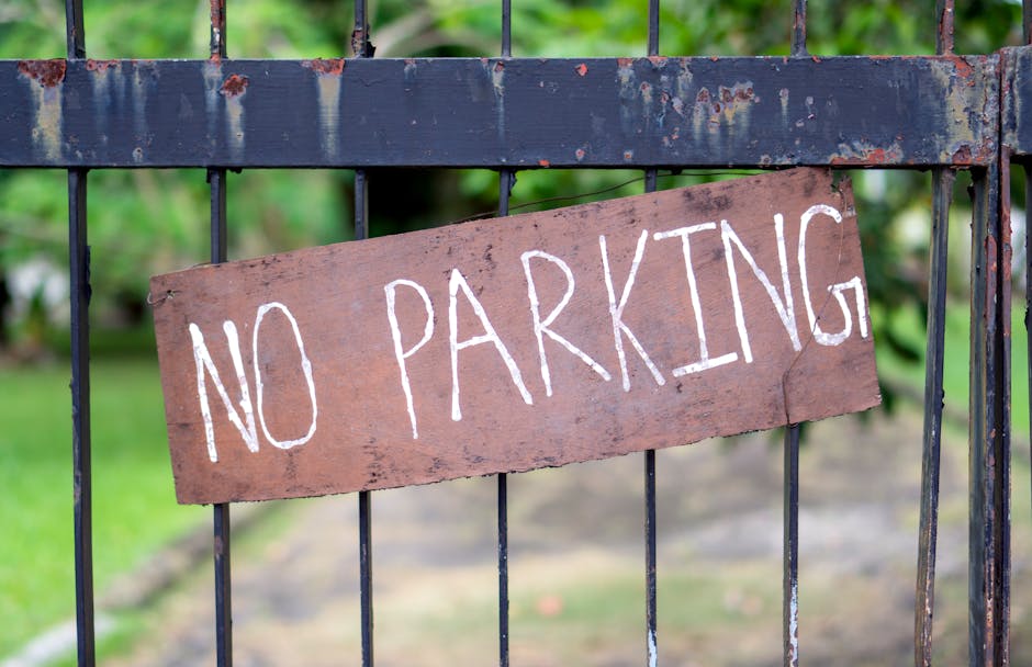 A close-up of a black metal gate with vertical bars, featuring a weathered wooden sign attached to it with visible rust and chipped paint. The sign displays the words 'NO PARKING' handwritten in white paint or chalk, with a slightly uneven appearance. Behind the gate, there is a blurred background of green foliage and a garden, suggesting the location is part of a residential property. The image is captured in natural daylight, emphasizing the texture of the metal and wood. This scene relates to house removals and the importance of navigating parking restrictions during relocation in Woodside Park, as managed by Man with Van Woodside Park.