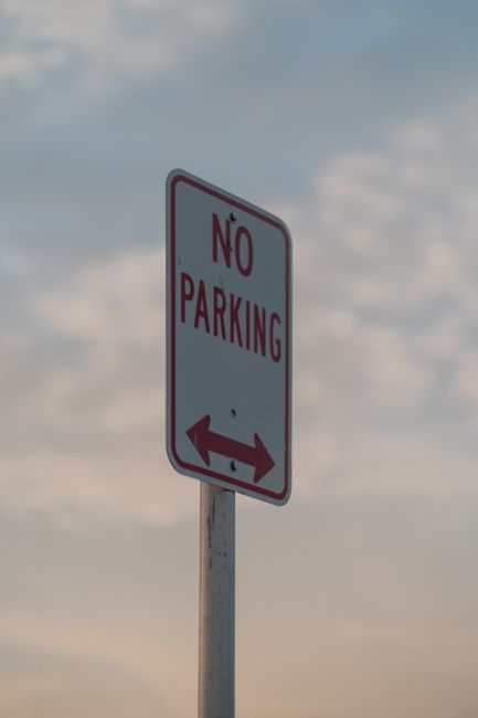 A tall, rectangular 'No Parking' sign with a white background and bold red and black lettering, mounted on a metal pole against a partly cloudy sky. The sign features a left and right arrow at the bottom indicating restricted parking in both directions. In the foreground, there are no visible vehicles or furniture, but the context suggests a street or driveway area near a house. The image is relevant to house removals and moving services, highlighting the importance of navigating parking restrictions during home relocation processes, as managed by companies like Man with Van Woodside Park.