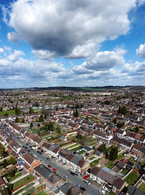 A view of a busy street in Woodside Park showing a row of multi-storey terraced houses with brick facades and pitched roofs, some with decorative gables. The street is lined with various small commerc