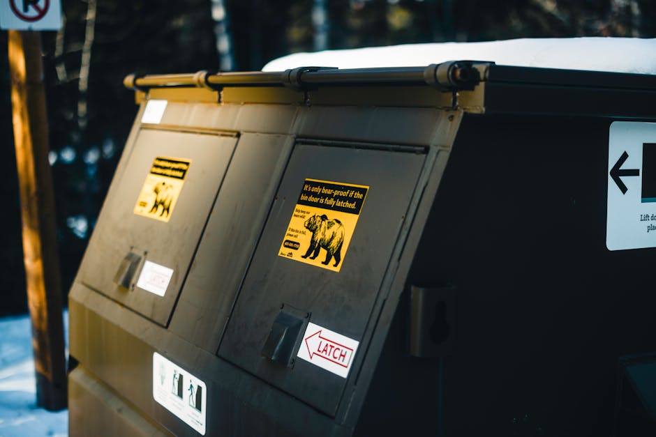 Close-up image of a large outdoor waste disposal bin with a black and yellow warning label indicating it is only bear-proof if fully latched, situated in an outdoor area with snow-covered ground and a wooden post in the background. The bin is made of metal with a sloped top, featuring multiple access compartments with small silver latches and white label stickers. The scene is illuminated by natural daylight, suggesting daytime. This kind of waste disposal unit is often used in bulk waste storage during home relocation processes, aligning with services like house removals and furniture transport. The image depicts a secure, outdoor waste management setup relevant to disposing of bulky waste before or after moving, and is associated with professional removals services like Man with Van Woodside Park. The environment and equipment shown illustrate proper waste handling in moving logistics contexts.