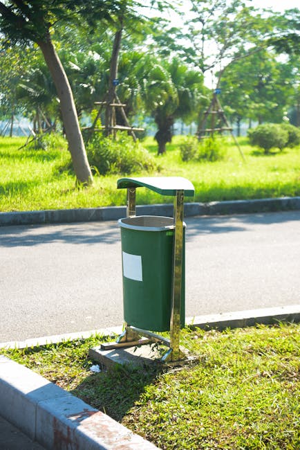 A green outdoor waste bin with a white label is mounted on a metal stand located on a grassy verge next to a paved road. The bin has a rounded top lid and appears to be made of plastic, with the metal support structure painted in yellow. In the background, there are lush green trees, including palm trees, and a park-like area with wooden scaffolding or support structures, along with shrubs and well-maintained greenery. The scene is illuminated by natural daylight, suggesting a bright, clear day. This setting depicts a typical urban or suburban roadside environment, potentially in a tropical or warm climate area, aligned with the context of household waste disposal and urban management, related to house removals or moving services such as those offered by Man with Van Woodside Park.