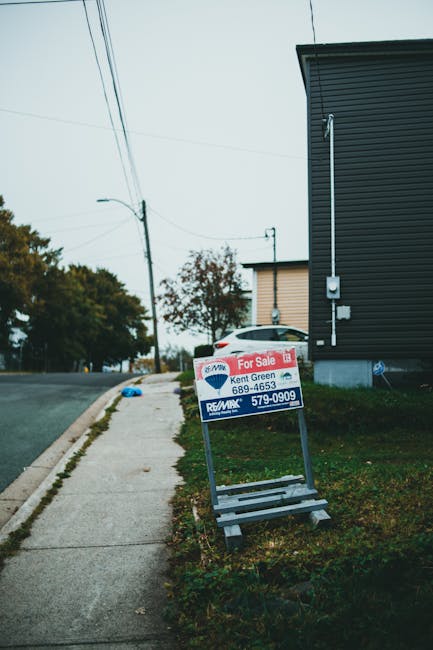 A metal A-frame signboard with a red and white 'For Sale' notice, attached to a metal frame with a blue and white 'RE/MAX' logo, positioned on a sidewalk next to a grassy area near a modern, dark-colored building with horizontal cladding. In the background, a white vehicle is partially visible, parked close to the building, with several trees and utility poles lining the street. Overhead power lines run along the road, which is slightly curved and free of traffic. The scene appears to be in a residential or commercial area during daylight, with natural lighting. This image illustrates the process of home relocation or property transition, as often handled by companies like Man with Van Woodside Park during moving and packing activities, including furniture transport and loading procedures, though no moving activity is visible in this specific shot.