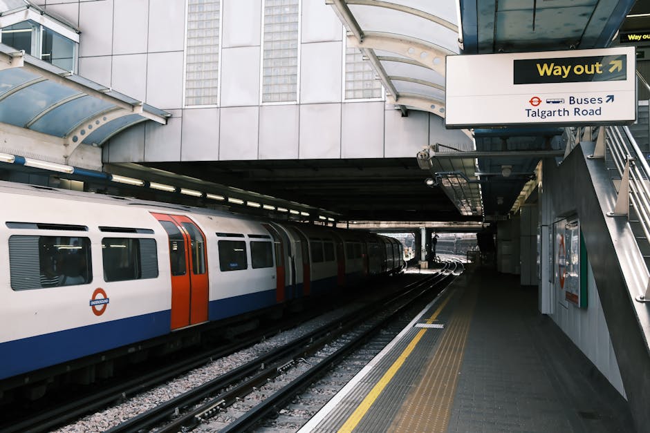 A view of a London Underground train with a white, orange, and blue exterior stationary at an outdoor station platform. The platform has a yellow tactile strip along the edge, with a grey tiled surface. Above, there is a curved glass and metal canopy providing shelter, and an overhead sign indicating directions to the bus stop on Talgarth Road and an outgoing route, with a yellow arrow and the London Underground roundel logo. To the right, a metal staircase leads up to an accessible pathway. The station appears modern, with a large building facade visible behind the canopy, and natural daylight illuminating the scene. This setting is relevant to house removals and packing logistics, illustrating temporary relocation points during furniture transport, which Man with Van Woodside Park offers in locations including Woodside Park and surrounding areas.