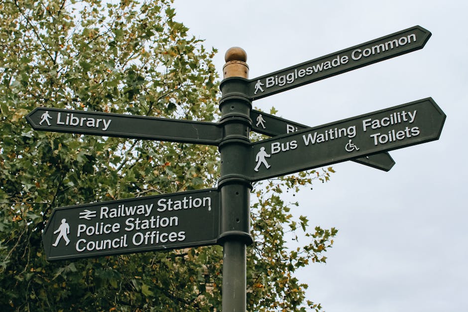 Photograph of a street signpost against a clear blue sky, featuring three directional signs. The top sign indicates Gillespie Park Nature Reserve, located 100 yards away, with a brown background and white text and icon of a duck. Below it, a black sign shows a pedestrian walking icon pointing toward Arsenal Football, with the words 'Arsenal Football' in white. Several stickers are attached to the signs, including one with orange and black design. The signpost is situated near leafy trees and residential buildings with chimneys, suggesting an urban or suburban environment. This setting relates to the context of house removals and moving services that Man with Van Woodside Park provides, emphasizing navigation and local area familiarisation during the relocation process.